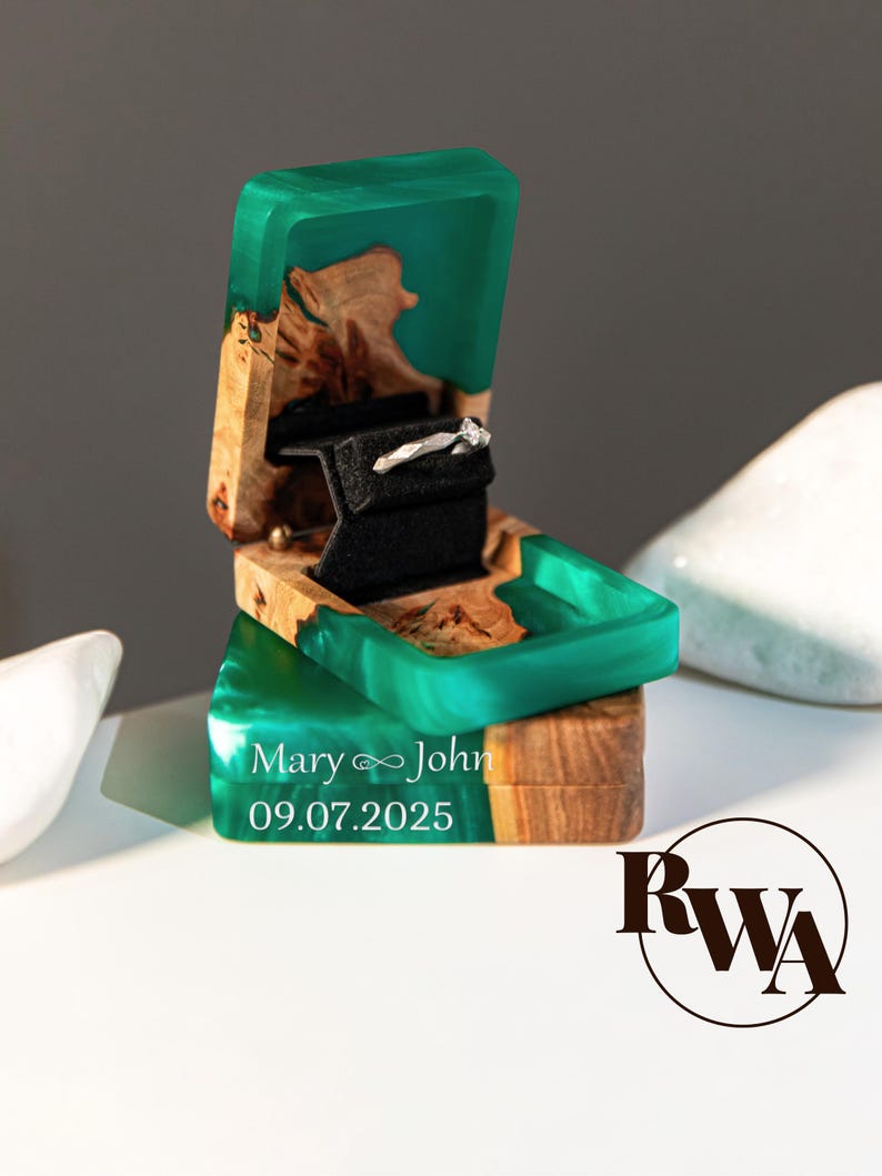 May include: A wooden ring box with a teal resin top and base, open to reveal a diamond ring on a black velvet insert. The box is engraved with the names "Mary &infin; John" and the date "09.07.2025".