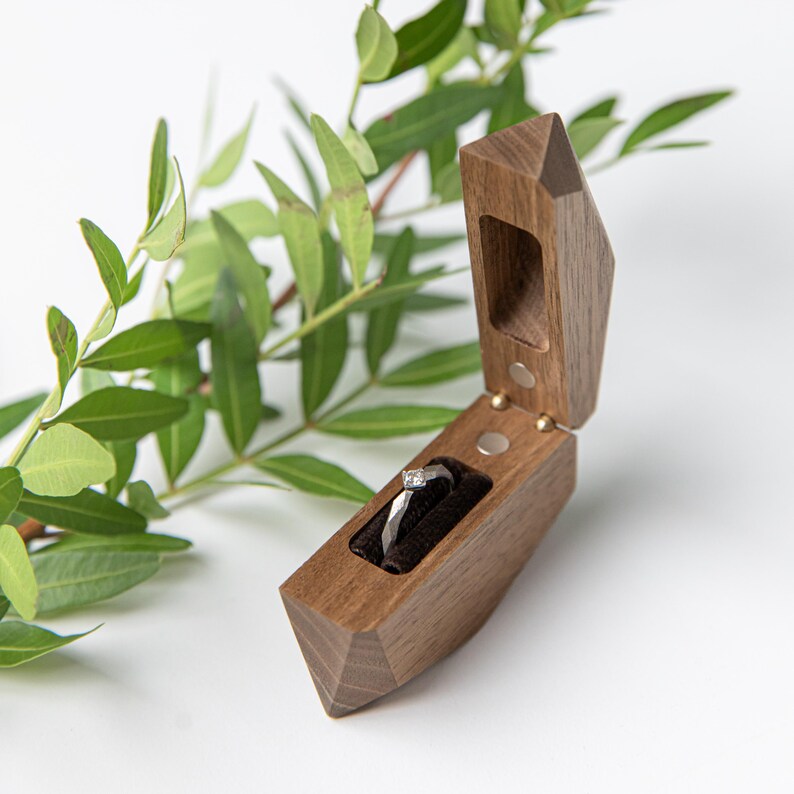 May include: A wooden ring box with a geometric design, open to reveal a diamond ring on a black velvet cushion. The box is made of dark wood and has a magnetic closure. Green leaves are in the background.