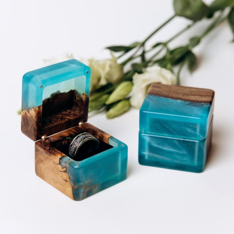 May include: Two square ring boxes crafted from natural wood and translucent turquoise resin. One box is open, revealing a silver-toned ring with an intricate engraved pattern inside, while the other box is closed, showcasing the smooth resin finish.
