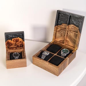 May include: Two wooden watch boxes with hinged lids featuring black resin accents and natural wood grain. The smaller box holds one wristwatch on a black cushion, while the larger box has four compartments with two wristwatches displayed.