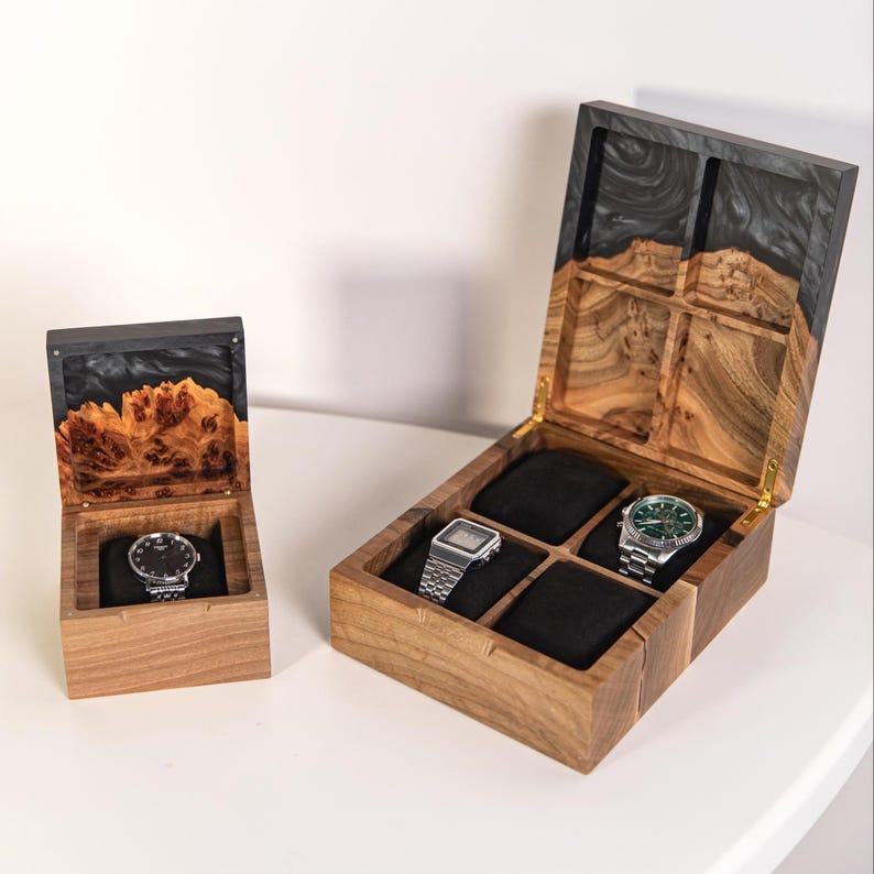 May include: Two wooden watch boxes with hinged lids featuring black resin accents and natural wood grain. The smaller box holds one wristwatch on a black cushion, while the larger box has four compartments with two wristwatches displayed.