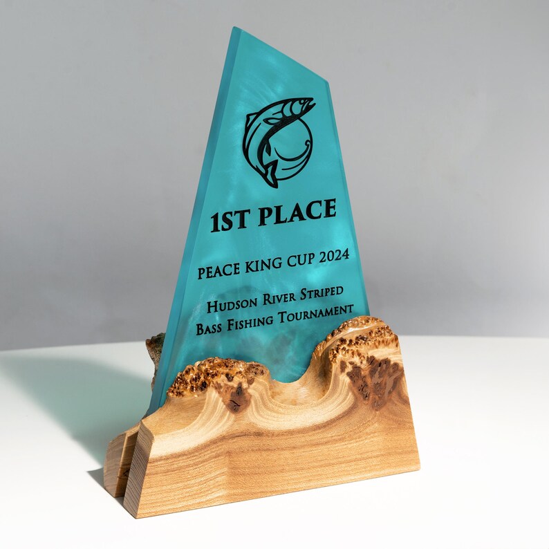 May include: A turquoise-colored trophy with a black fish logo and the text "1ST PLACE". The trophy is for the "Peace King Cup 2024 Hudson River Striped Bass Fishing Tournament". It is mounted on a wooden base.