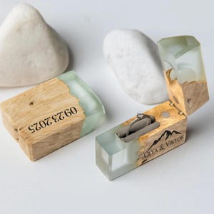 May include: Wooden ring box with a clear, light green resin top and a silver ring inside. The box is engraved with the date "09.23.2025" and the names "OLYA & VIKTOR" with a mountain graphic. The box is made of light brown wood.
