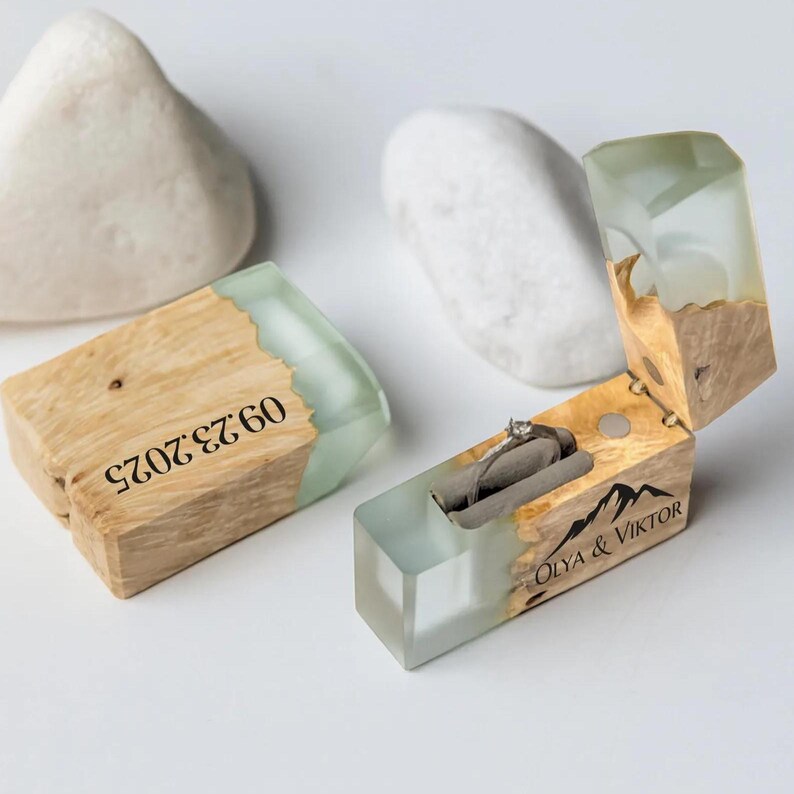 May include: Wooden ring box with a clear, light green resin top and a silver ring inside. The box is engraved with the date "09.23.2025" and the names "OLYA & VIKTOR" with a mountain graphic. The box is made of light brown wood.