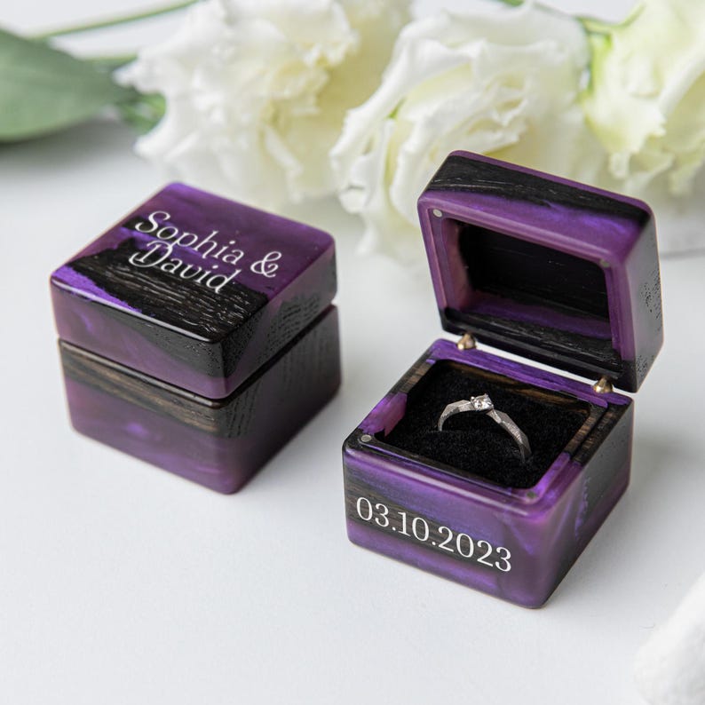 May include: Two square ring boxes made of dark wood and purple resin. One box is closed with the text Sophia & David engraved on top. The other box is open, revealing a silver ring with a small stone inside, with the date 03.10.2023 on the front.