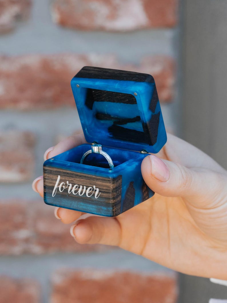 May include: A square ring box crafted from dark wood and vibrant blue resin, featuring a hinged lid and the word forever engraved in white script on the front. Inside the box sits a thin silver ring with a small blue stone setting.