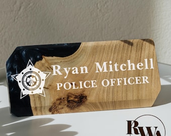 May include: A rectangular desk nameplate made of natural wood and dark blue resin. The wood features a prominent grain and knot. White text reads Ryan Mitchell and POLICE OFFICER, accompanied by a white star-shaped police badge graphic.