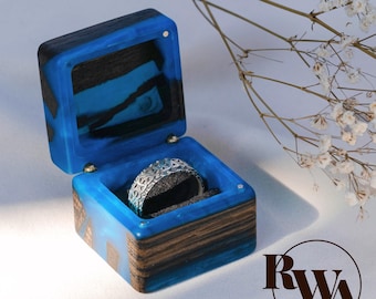 May include: A square ring box crafted from dark wood and vibrant blue resin, featuring a hinged lid and a silver-toned metal ring with an intricate filigree pattern resting inside on a dark grey velvet insert. The letters RWA are visible.