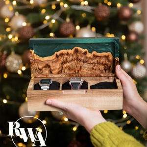 May include: A rectangular wooden watch box with three interior compartments and black velvet cushions. The lid features a natural wood grain edge combined with a smooth, dark green resin inlay. Two wristwatches are stored inside the box.