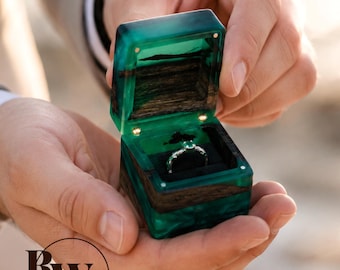 Könnte beinhalten: Eine quadratische Ringbox aus dunklem Holz und durchscheinendem smaragdgrünem Harz mit aufklappbarem Deckel und schwarzem Innenfutter. Im Inneren befindet sich ein silberner Ring mit einem kleinen grünen Edelstein. Die Buchstaben RWA sind in Schwarz aufgedruckt.