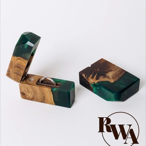 Può includere: Due scatole rettangolari per anelli realizzate in legno naturale e resina verde smeraldo traslucida. Una scatola è aperta, rivelando un interno in velluto marrone scuro che contiene un anello d'argento con una piccola pietra. L'altra è chiusa. Il logo RWA è visibile.