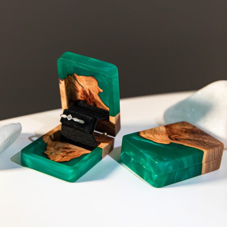 Op de afbeelding: Een rechthoekig ringdoosje gemaakt van natuurlijk hout en doorschijnende smaragdgroene hars. Het open doosje heeft een zwarte fluwelen inzet met een zilveren ring met een kleine vierkante steen. Een tweede bijpassend doosje staat ernaast.