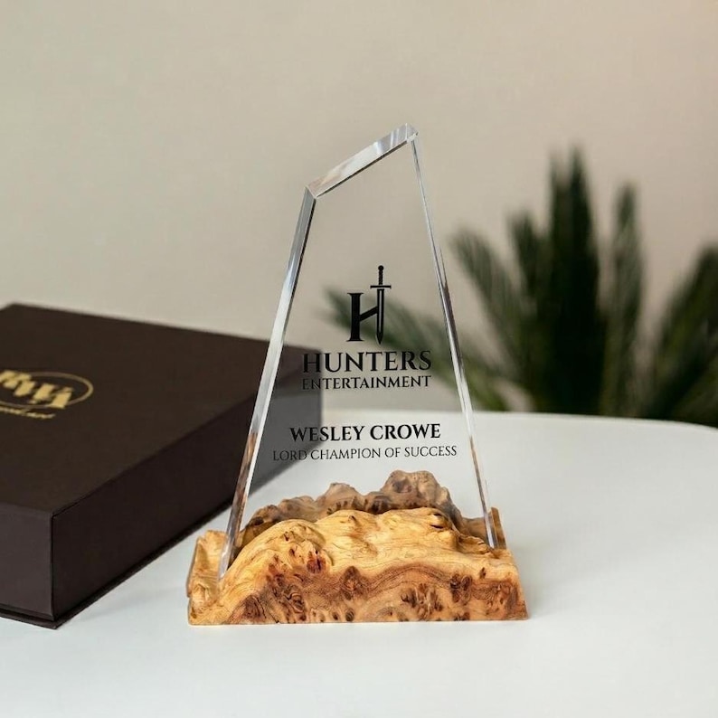 May include: A clear glass award with a wooden base. The award has the text "Hunters Entertainment" and "Wesley Crowe Lord Champion of Success". The wooden base has a natural, textured appearance.