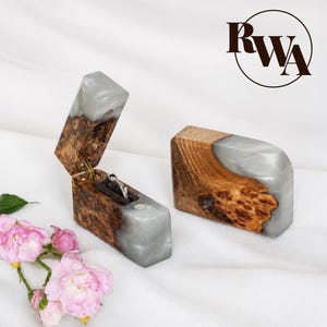 Può includere: Due scatole rettangolari per anelli realizzate in legno di radica naturale e resina grigia traslucida. Una scatola è aperta, rivelando un inserto in velluto nero che contiene un anello d'argento, mentre l'altra è chiusa. Il logo RWA è visibile nell'angolo in alto a destra.