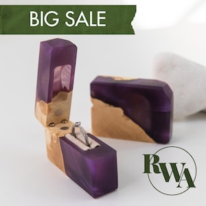 Può includere: Un portagioie rettangolare in resina viola e legno chiaro, con il coperchio aperto che contiene un anello argentato. Il testo BIG SALE è visualizzato su un banner verde in alto e il logo RWA è visibile nell'angolo in basso a destra.
