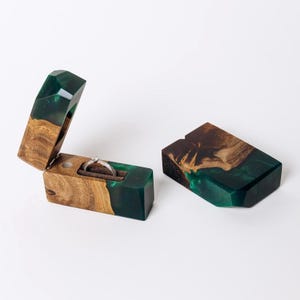 May include: A wooden ring box with a hinged lid, open to reveal a silver ring. The box is made of wood and dark green resin. The closed box is also visible. The ring box is on a white background.