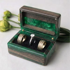 May include: A rectangular ring box crafted from dark wood and translucent emerald green resin. The interior features a black velvet insert holding two wide metal bands with intricate patterns and one central solitaire ring with a clear stone.