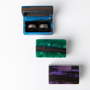 May include: Three rectangular ring boxes made of dark wood and colored resin. One box is open, revealing two silver-toned patterned rings inside. The other two boxes are closed, featuring swirling green and purple resin accents respectively.
