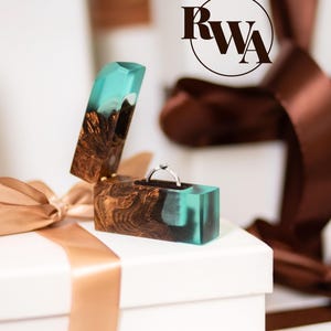 Op de afbeelding: Een rechthoekig ringdoosje gemaakt van donkerbruin hout en doorschijnende turquoise hars, met een scharnierend deksel en een zilveren ring met een kleine steen erin. Het logo RWA is zichtbaar in de rechterbovenhoek van de afbeelding.