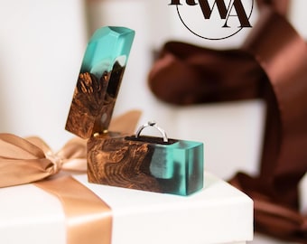 Op de afbeelding: Een rechthoekig ringdoosje gemaakt van donkerbruin hout en doorschijnende turquoise hars, met een scharnierend deksel en een zilveren ring met een kleine steen erin. Het logo RWA is zichtbaar in de rechterbovenhoek van de afbeelding.