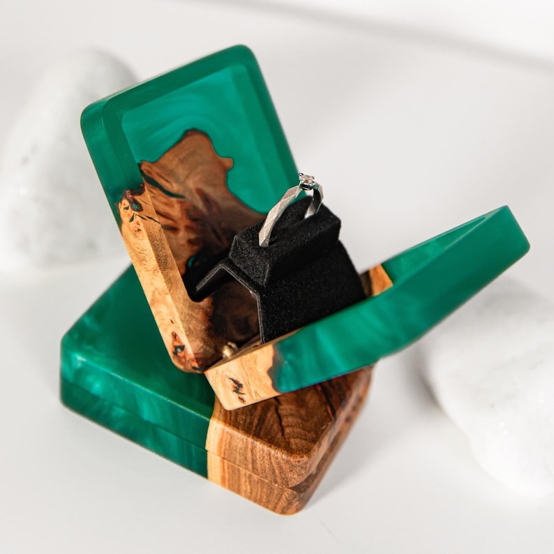 Op de afbeelding: Een ringdoosje gemaakt van natuurlijk hout en doorschijnende smaragdgroene hars, voorzien van een scharnierend deksel en een zwart fluwelen inzetstuk met een zilverkleurige ring met een kleine heldere steen. Het doosje heeft een rechthoekige basis.