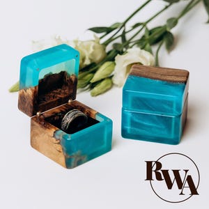 May include: A handcrafted ring box with a unique design. The box is made of wood and resin, with a vibrant turquoise resin interior and exterior. The box is open, revealing a silver ring inside. The box is square and has a hinged lid.