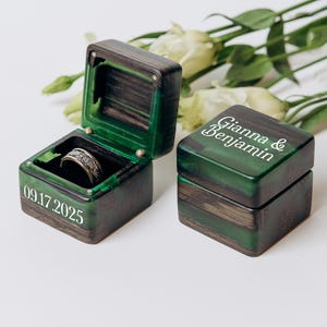 May include: Two square ring boxes made of dark wood and green resin. One box is open, revealing a silver-toned patterned ring inside, with the date 09.17.2025 printed on the side. The closed box features the names Gianna & Benjamin in white text.