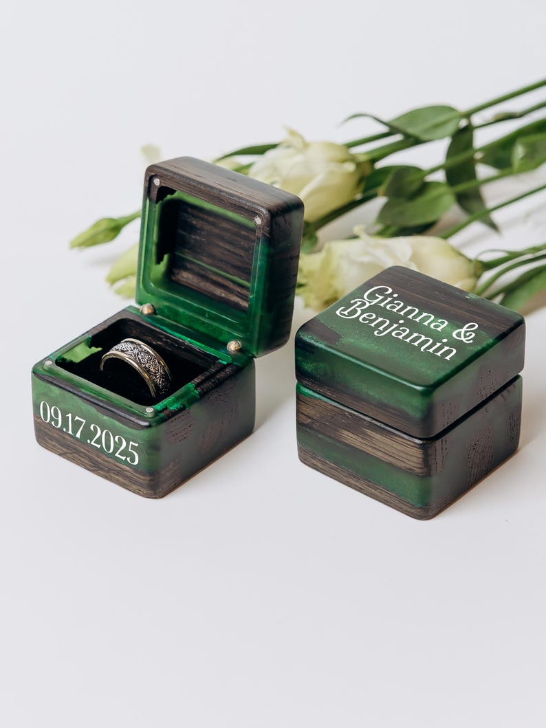 May include: Two square ring boxes made of dark wood and green resin. One box is open, revealing a silver-toned patterned ring inside, with the date 09.17.2025 printed on the side. The closed box features the names Gianna & Benjamin in white text.