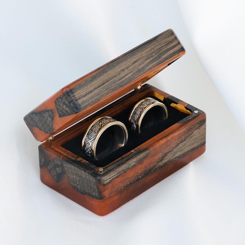 May include: A rectangular ring box crafted from dark wood and translucent amber-colored resin, featuring a hinged lid. Inside, two silver-toned metal rings with intricate, dark engraved patterns are nestled within a black velvet-lined interior.