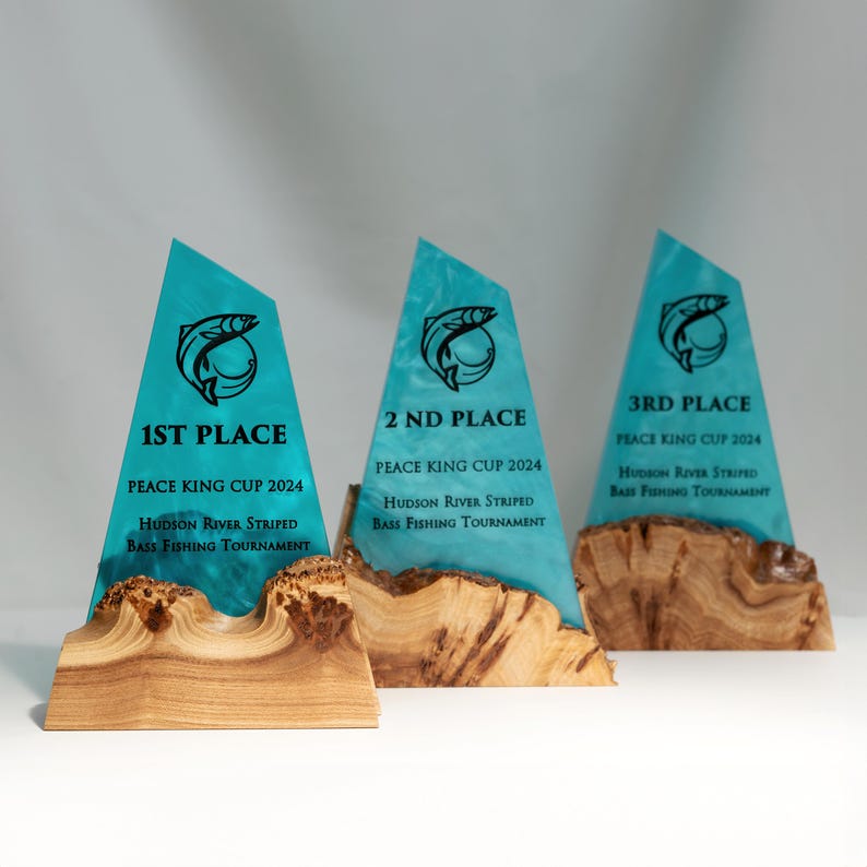 May include: Three fishing tournament trophies. Each trophy has a teal-colored, triangular top with a fish graphic and text indicating placement: 1st, 2nd, and 3rd place. The bases are made of wood, and the text includes the tournament name and year.