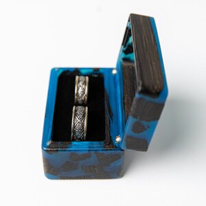 May include: A rectangular ring box crafted from dark wood and vibrant blue resin, featuring a hinged lid and a black velvet interior. Two silver-toned metal rings with intricate, embossed scrollwork patterns are positioned upright inside the box.