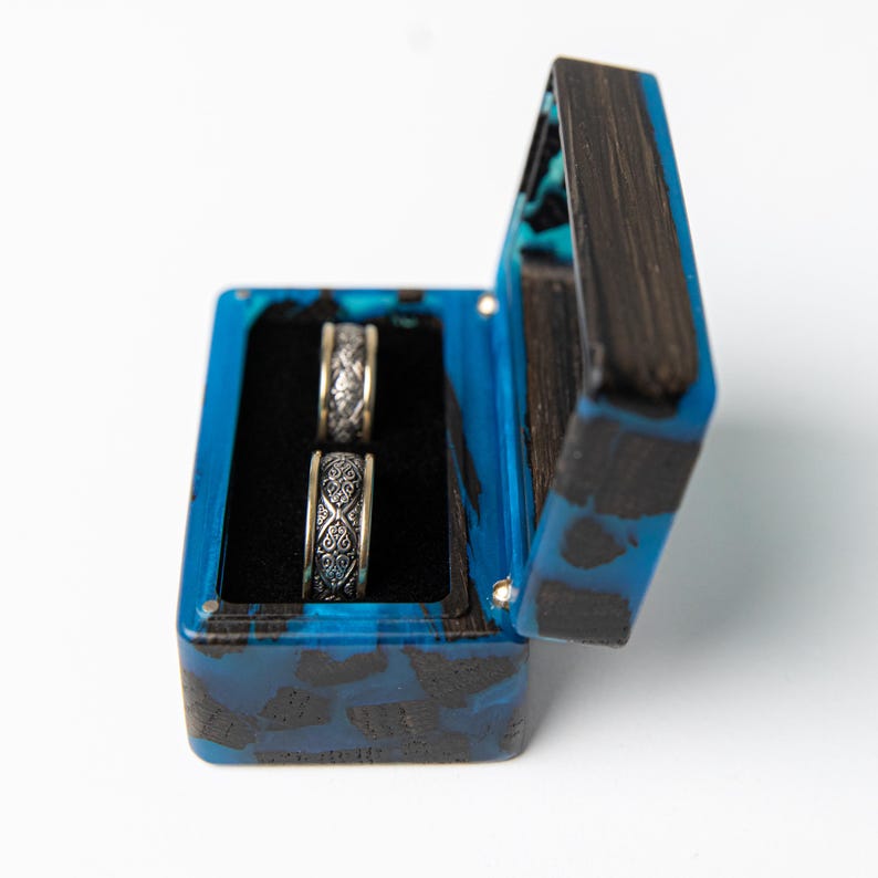 May include: A rectangular ring box crafted from dark wood and vibrant blue resin, featuring a hinged lid and a black velvet interior. Two silver-toned metal rings with intricate, embossed scrollwork patterns are positioned upright inside the box.