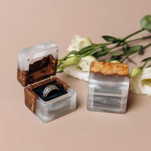 May include: Two square ring boxes crafted from natural wood and translucent white resin. One box is open, revealing a dark velvet interior holding a silver-toned band with an intricate pattern. The other box is closed, showing the wood and resin.