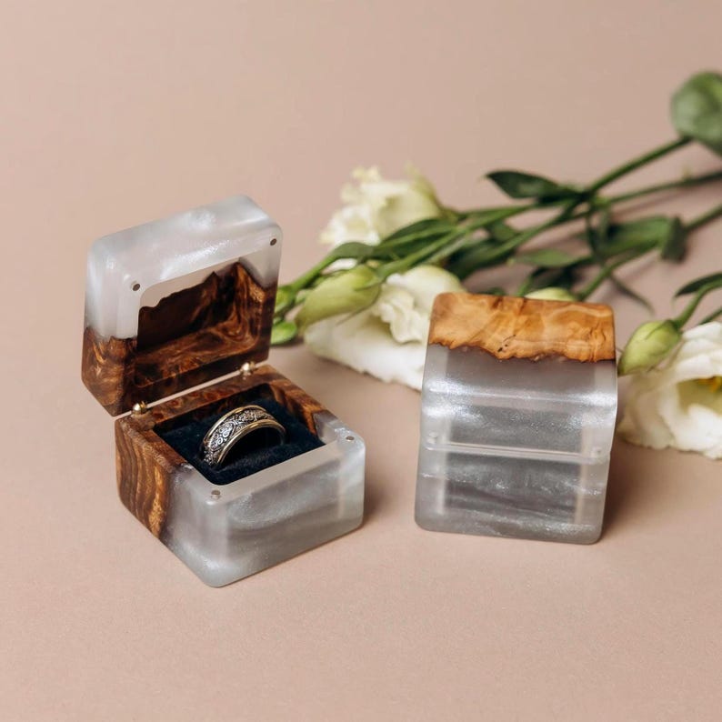 May include: Two square ring boxes crafted from natural wood and translucent white resin. One box is open, revealing a dark velvet interior holding a silver-toned band with an intricate pattern. The other box is closed, showing the wood and resin.
