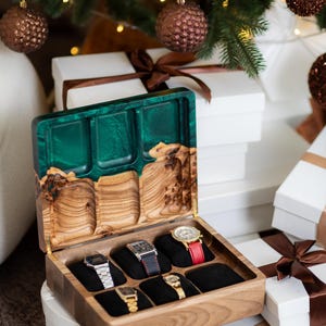 May include: A wooden watch storage box with a hinged lid featuring a green resin inlay. The interior contains six black velvet cushions, five of which hold wristwatches with various metal and leather bands. The lid interior has three carved wooden compartments.