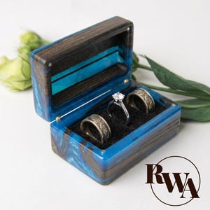 May include: A rectangular ring box crafted from dark wood and vibrant blue resin, featuring a hinged lid. The interior is lined with black fabric and holds two wide, patterned metal bands and one silver-toned ring with a central clear stone.