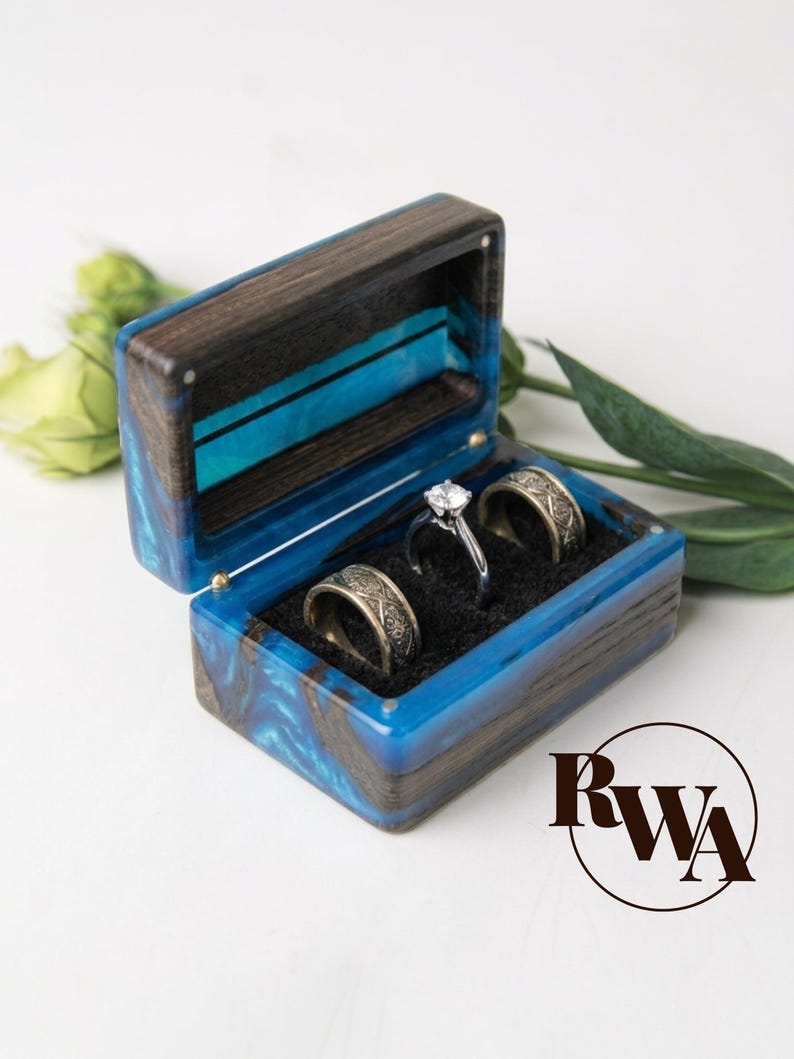May include: A rectangular ring box crafted from dark wood and vibrant blue resin, featuring a hinged lid. The interior is lined with black fabric and holds two wide, patterned metal bands and one silver-toned ring with a central clear stone.