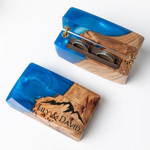 May include: A wooden ring box with a blue resin top and a mountain design. The box is open, revealing two rings with intricate patterns. The text "LILY & DAVID" is visible on the closed box. The box is made of wood and resin.
