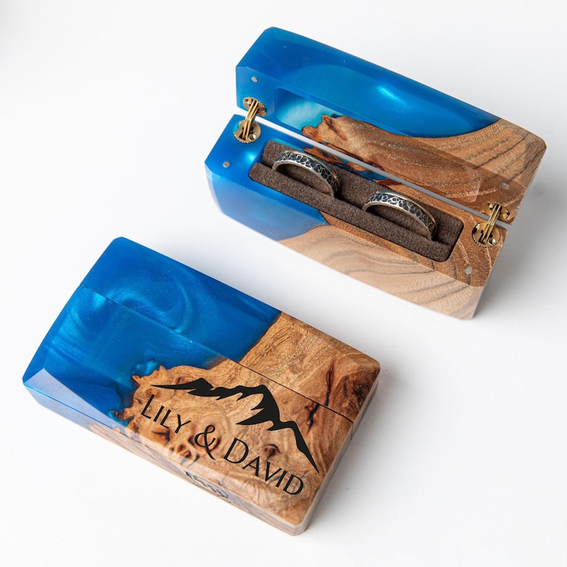 May include: A wooden ring box with a blue resin top and a mountain design. The box is open, revealing two rings with intricate patterns. The text "LILY & DAVID" is visible on the closed box. The box is made of wood and resin.