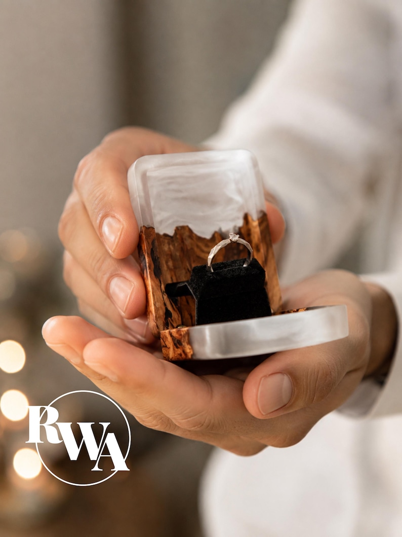 Op de afbeelding: Een kleine rechthoekige ringdoos gemaakt van een combinatie van natuurlijk bruin hout en doorschijnende witte hars. De binnenkant is voorzien van een zwart fluwelen inzetstuk met een dunne zilverkleurige ring met een kleine heldere steen.