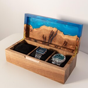 May include: A rectangular wooden watch box with a hinged lid featuring a blue resin inlay. The interior is divided into three compartments with black velvet cushions, holding a digital watch and an analog watch with a green face.