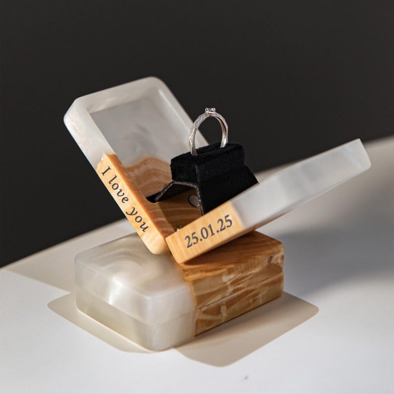 May include: A wooden ring box with a silver ring on a black velvet stand. The box is open, revealing the ring. The box has a white and wood grain finish with the text "I love you" and "25.01.25" engraved on it.