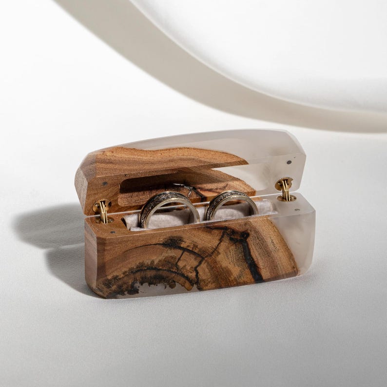May include: A wooden ring box with a clear resin top, showcasing two silver rings. The box is open, revealing the rings nestled on a soft, white interior. The wood grain is visible, adding a natural touch.