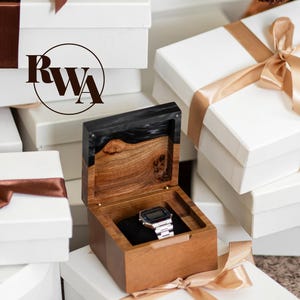 May include: A square wooden watch box with a hinged lid featuring a black resin accent and a natural wood grain finish. Inside, a silver digital watch with a metal link band rests on a black velvet cushion. The logo RWA is visible.