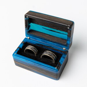 May include: A rectangular ring box crafted from dark wood and vibrant blue resin, featuring a hinged lid and a black velvet interior. Two silver-toned metal rings with intricate, embossed patterns are nestled inside the box on the black lining.