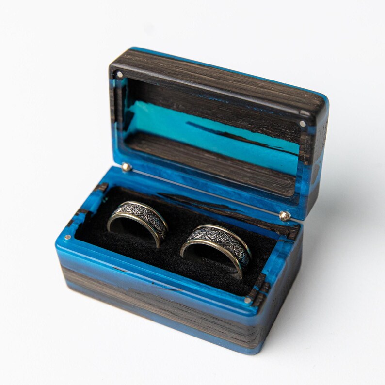 May include: A rectangular ring box crafted from dark wood and vibrant blue resin, featuring a hinged lid and a black velvet interior. Two silver-toned metal rings with intricate, embossed patterns are nestled inside the box on the black lining.