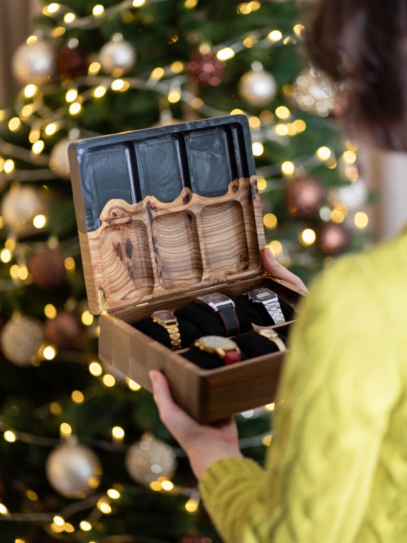 May include: A rectangular wooden watch box with a hinged lid featuring a dark resin top section and a natural wood grain base. The interior contains six individual compartments, each fitted with a black velvet cushion holding a wristwatch.