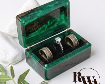 May include: A rectangular ring box crafted from dark wood and translucent emerald green resin. The open lid reveals a black interior lining holding two wide, engraved metal bands and one central silver ring with a clear round stone.