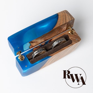May include: Open wooden ring box with a blue resin interior and two rings. The box is made of wood and blue resin, with gold-colored hinges. The rings are silver with a textured design, set in a brown velvet interior.