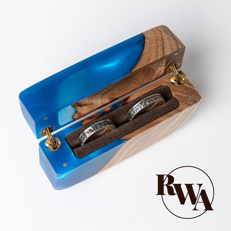 May include: Open wooden ring box with a blue resin interior and two rings. The box is made of wood and blue resin, with gold-colored hinges. The rings are silver with a textured design, set in a brown velvet interior.